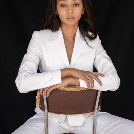 Synclaire Warren wears a white suit and sits on a backwards chair against an all black background