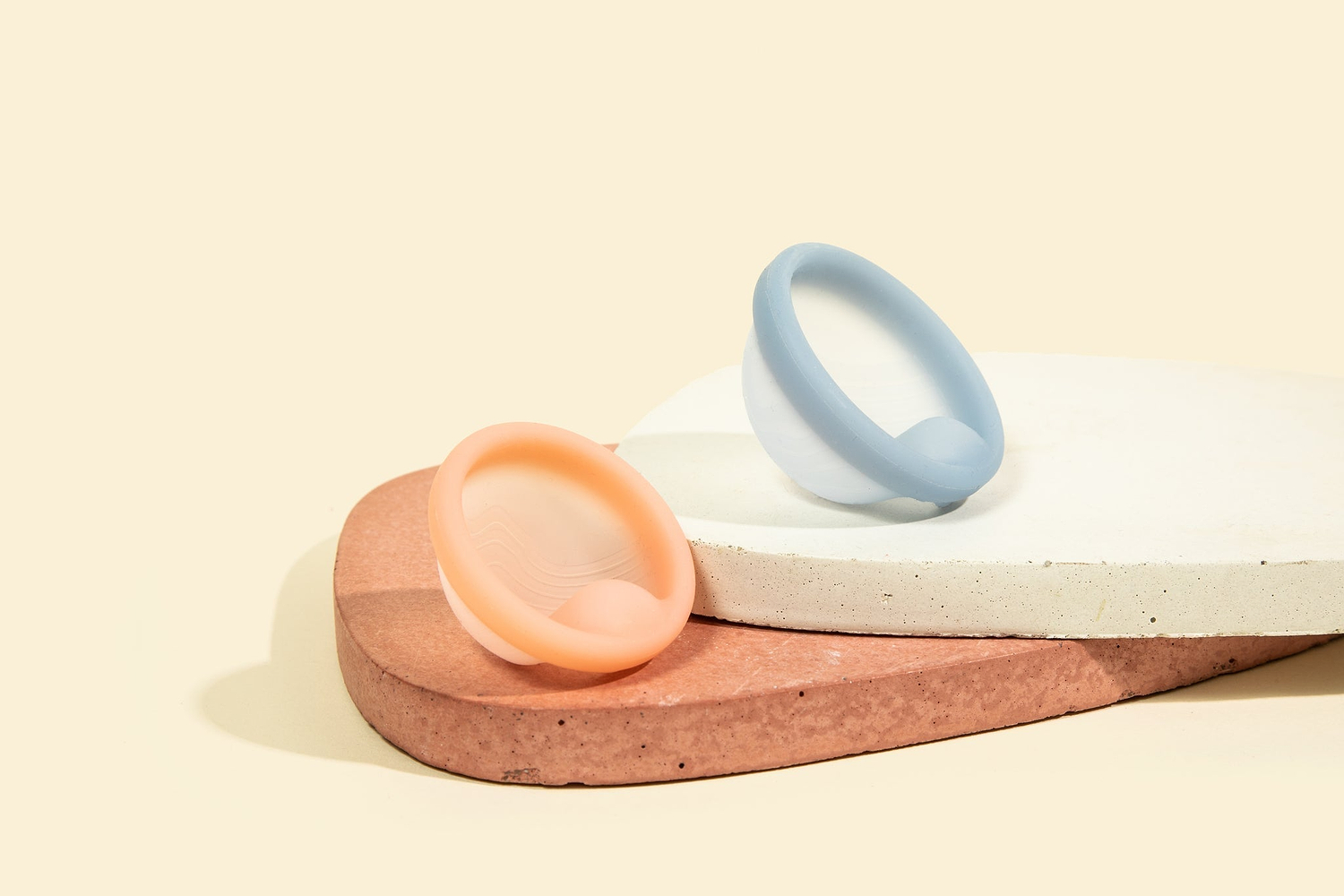 Two pastel menstrual discs displayed on stacked stone platforms against a beige background.
