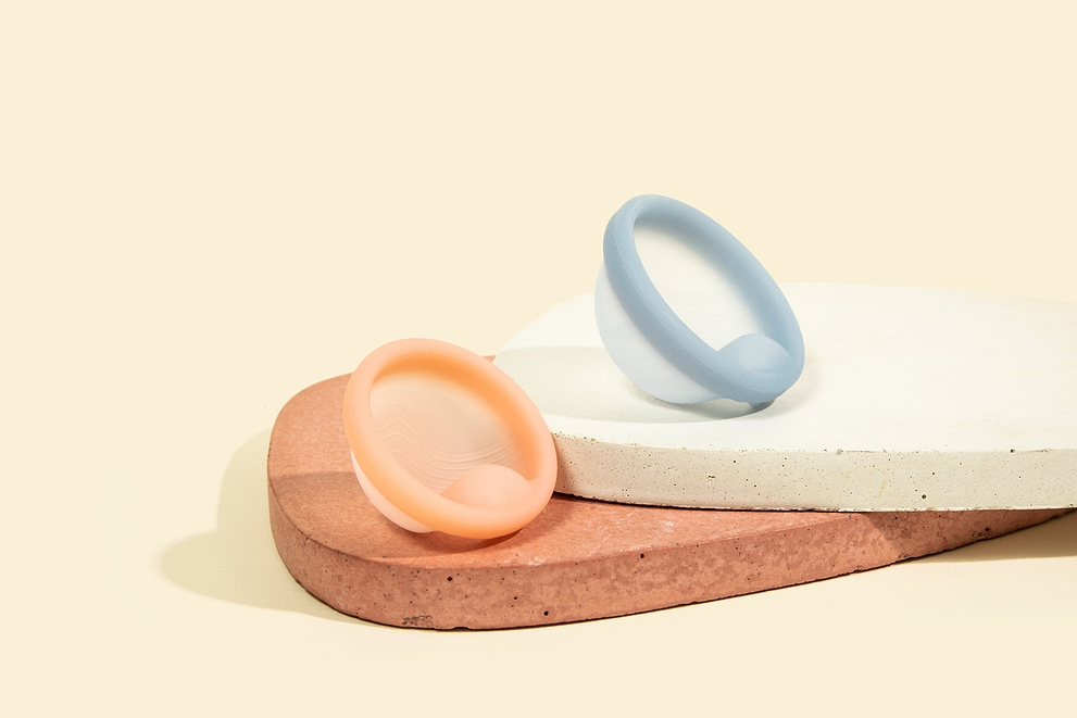 Two pastel menstrual discs displayed on stacked stone platforms against a beige background.