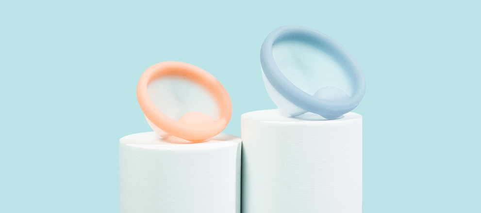 Peach and blue menstrual discs displayed on white pedestals against a soft blue background.