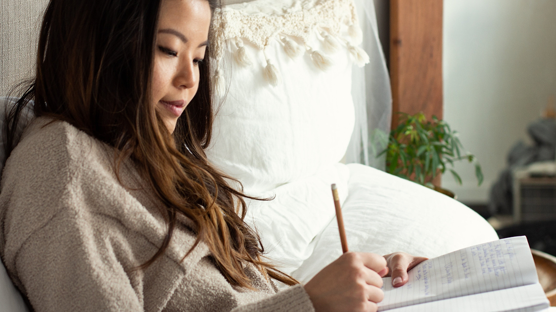 Woman in cozy loungewear writing in a notebook while sitting on a bed in a softly lit room.