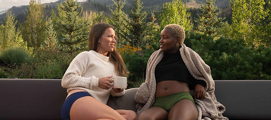 Two women relaxing outdoors in underwear, chatting with a mountain view behind them.