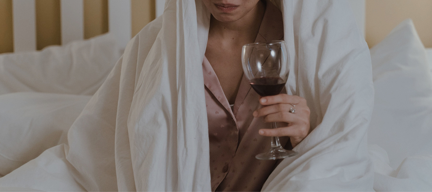 Person wrapped in a white blanket holding a glass of red wine while sitting on a bed.