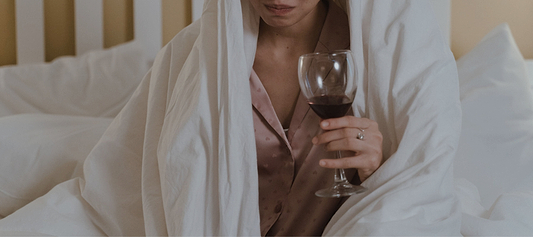Person wrapped in a white blanket holding a glass of red wine while sitting on a bed.