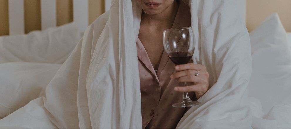 Person wrapped in a white blanket holding a glass of red wine while sitting on a bed.