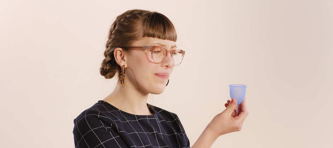 Woman with braided hair and glasses holding a blue menstrual cup against a neutral background.