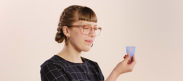 Woman with braided hair and glasses holding a blue menstrual cup against a neutral background.