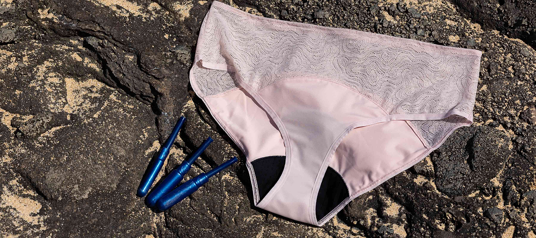 Light pink period underwear and blue applicators displayed on dark volcanic rocks outdoors.