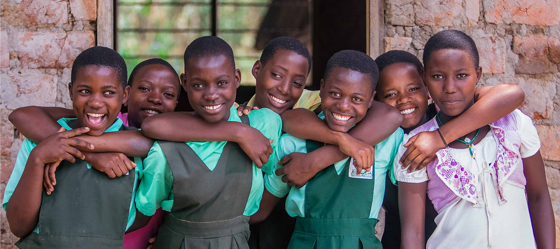 Seven smiling schoolgirls in uniforms standing together outside a brick building with arms around each other.
