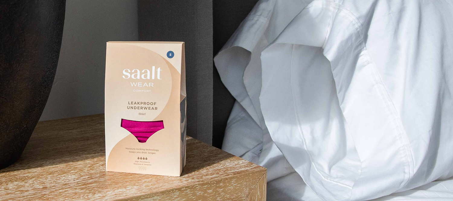 Box of leakproof underwear on a bedside table beside white bedding and dark curtains.