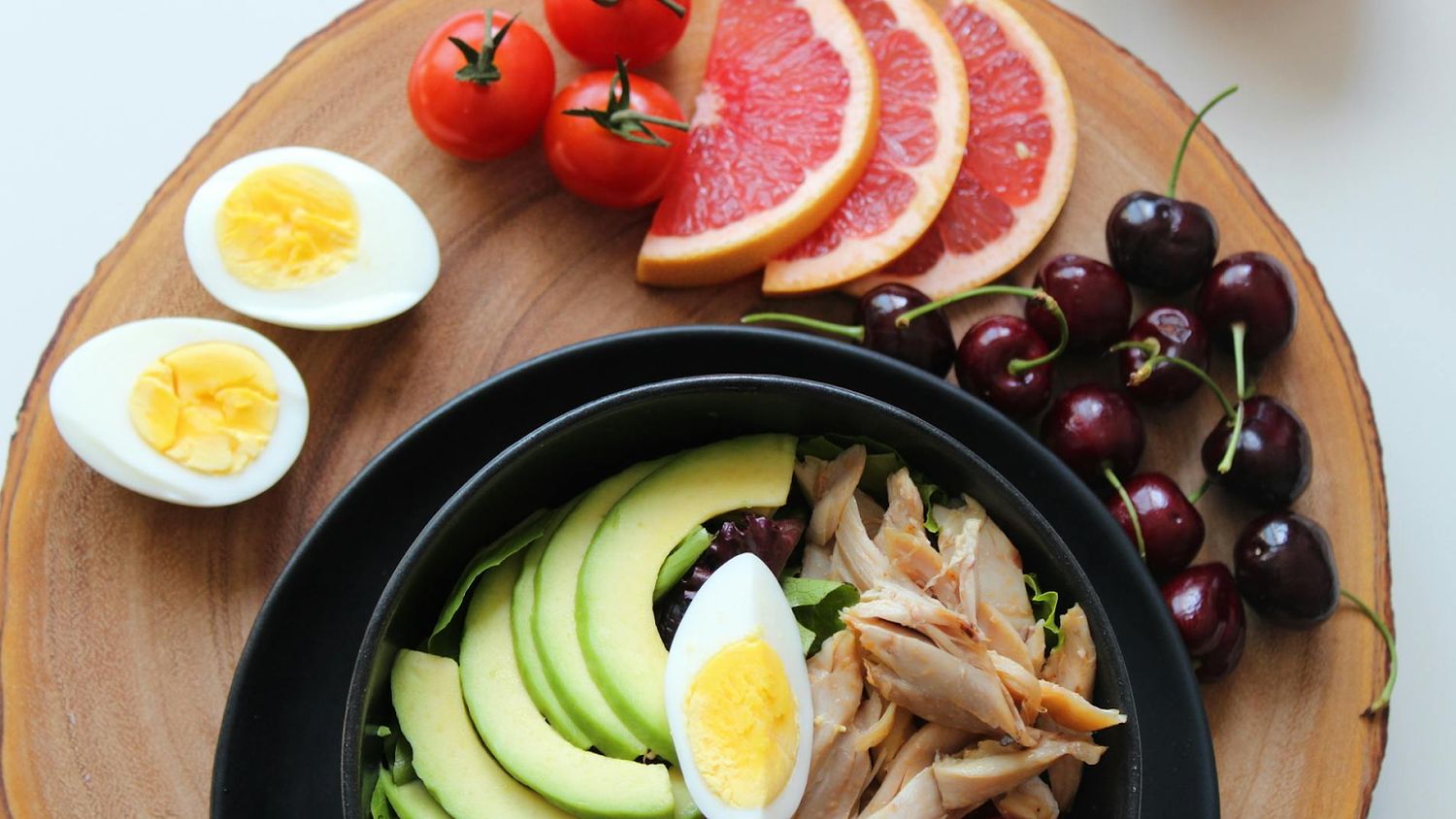 Healthy meal bowl with avocado, egg, and chicken, served with grapefruit, cherries, tomatoes, and eggs.