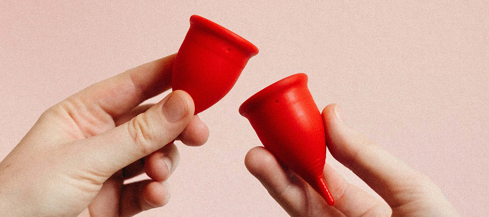 Two hands holding red menstrual cups against a soft pink background.
