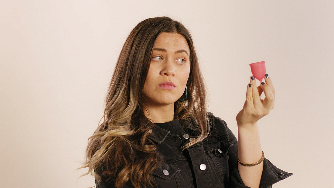 Woman holding and examining a pink menstrual cup against a neutral background.