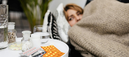 Person resting under a blanket beside a table with medicine bottles, pills, and glasses of water.