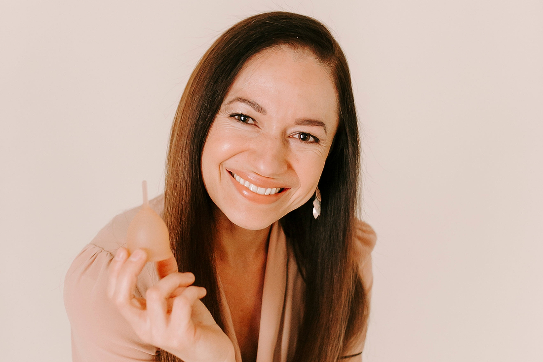 Smiling woman holding a beige menstrual cup against a soft neutral background.