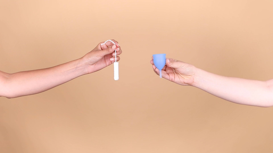 Two hands holding menstrual care products, comparing a tampon and a reusable menstrual cup against a neutral background.