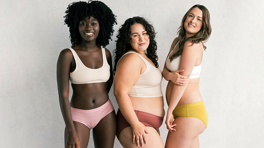 Three diverse women smiling in neutral underwear and bras, posing together against a light studio backdrop.