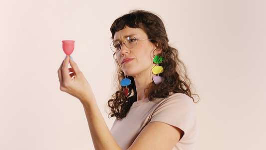 Woman with colorful earrings examining a pink menstrual cup against a plain background.