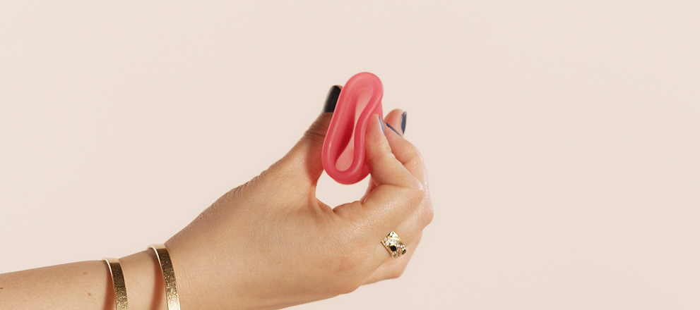 Hand holding a pink menstrual cup, wearing rings and bracelets, against a neutral background