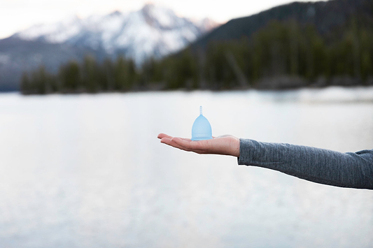 Hand holding a blue menstrual cup outdoors with snowy mountains and pine trees in the background