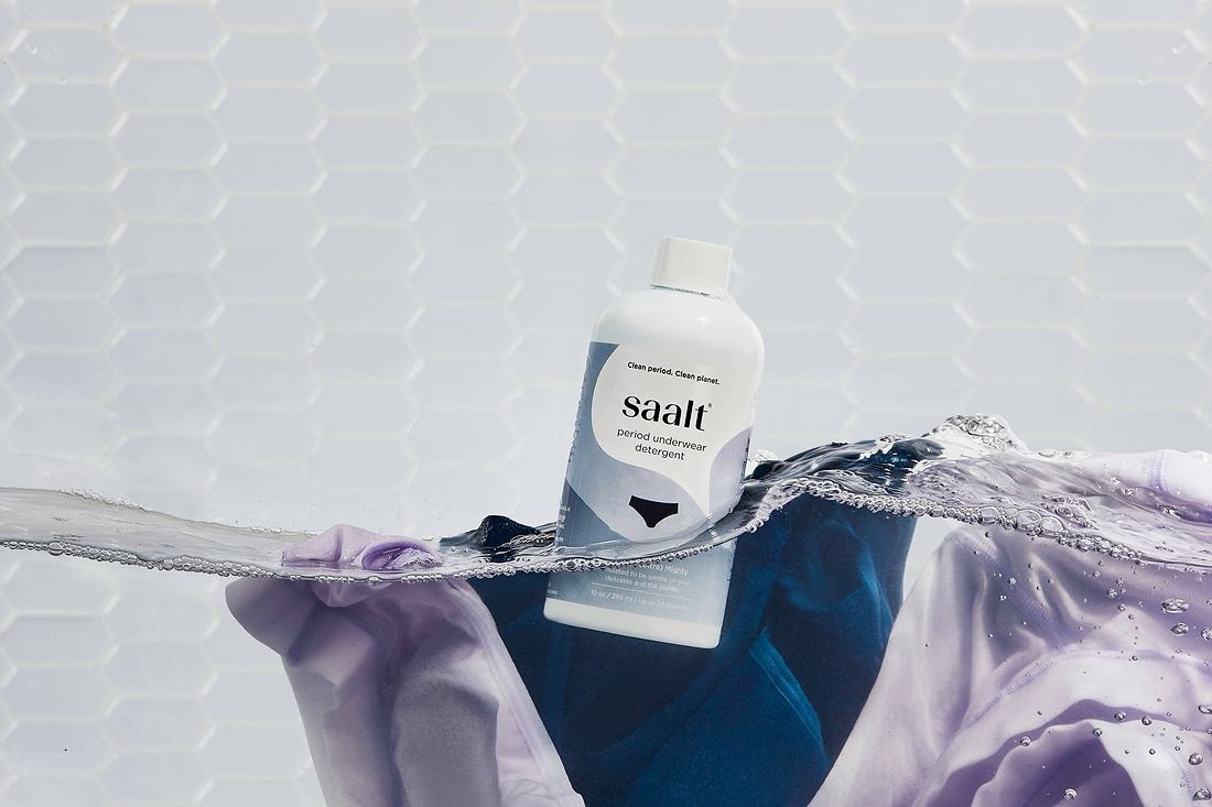 Saalt period underwear detergent bottle floating in water with purple fabric against a white tiled background