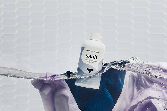 Saalt period underwear detergent bottle floating in water with purple fabric against a white tiled background