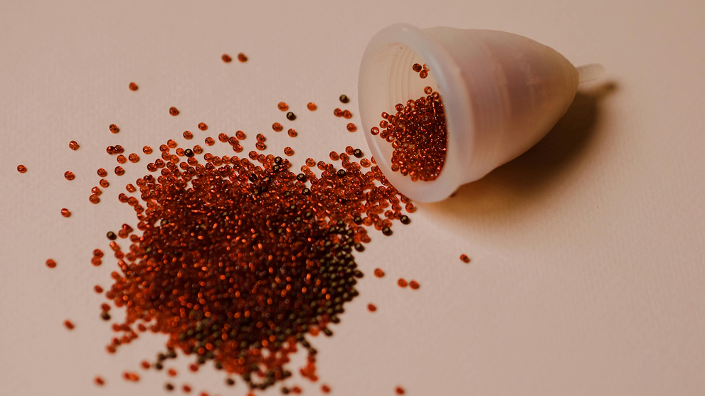 Menstrual cup tipped over with red beads symbolizing menstrual blood on a soft pink background.