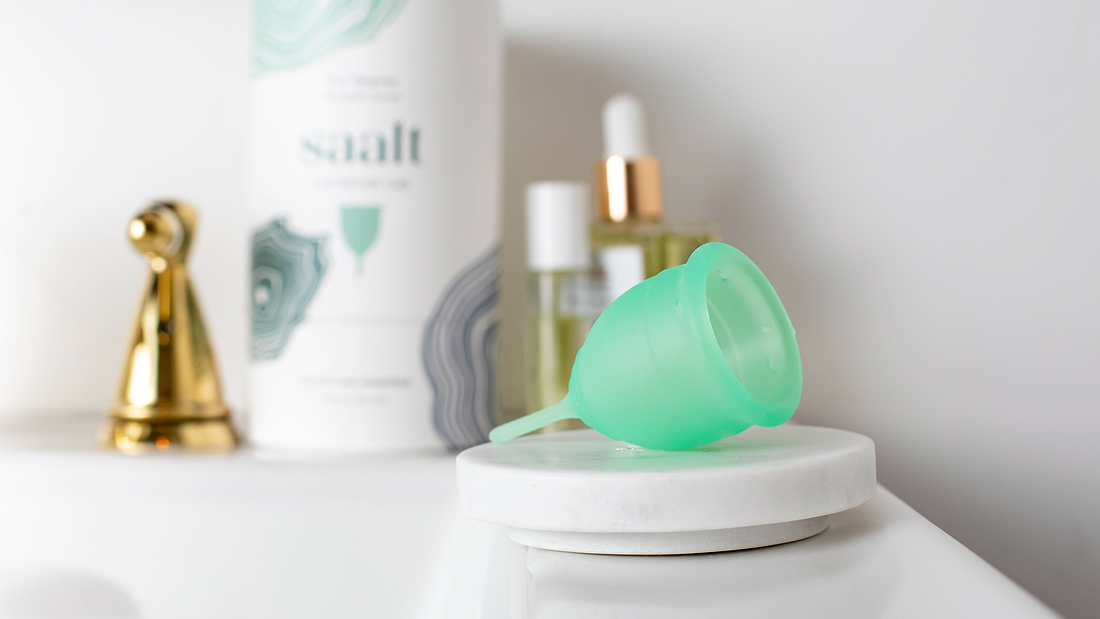 Green menstrual cup on bathroom counter beside toiletries and Saalt packaging.
