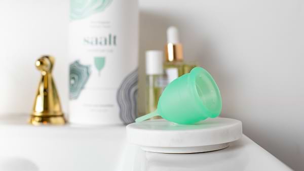 Green menstrual cup on bathroom counter beside toiletries and Saalt packaging.