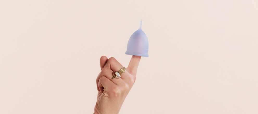 Hand balancing a lavender menstrual cup on one finger against a plain background.