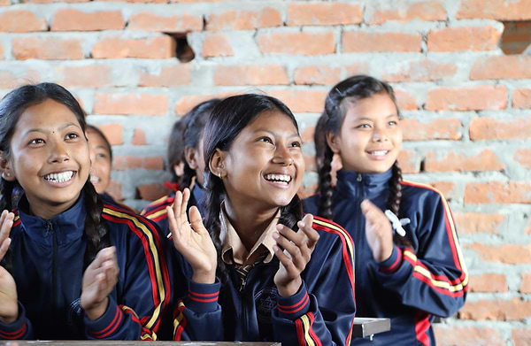 Smiling schoolgirls in matching uniforms clap together in a classroom with exposed brick walls.