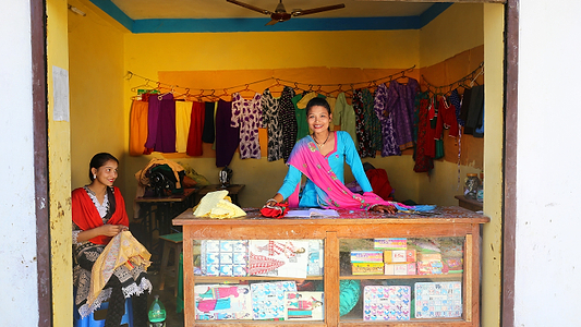 Two women smiling inside a colorful clothing and tailoring shop with garments hanging along the walls.