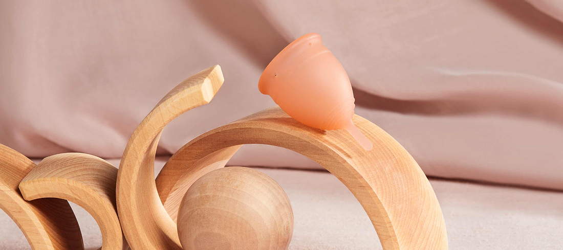 Peach-colored menstrual cup balanced on curved wooden toys against a soft pink background.