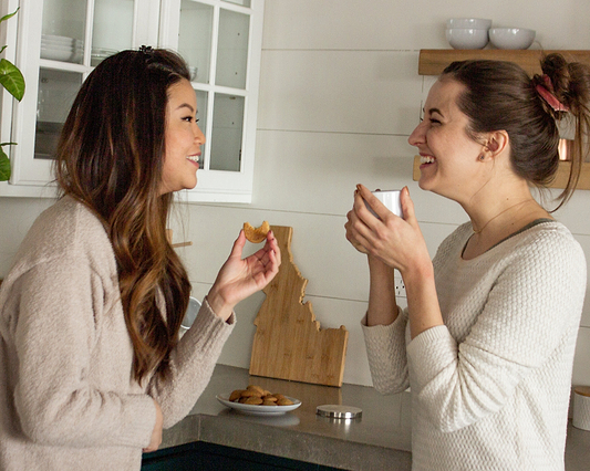 Two people laughing together in a kitchen while sharing cookies and drinks.