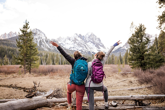 Two hikers with backpacks celebrate in front of snowy mountains and pine trees on a scenic trail.