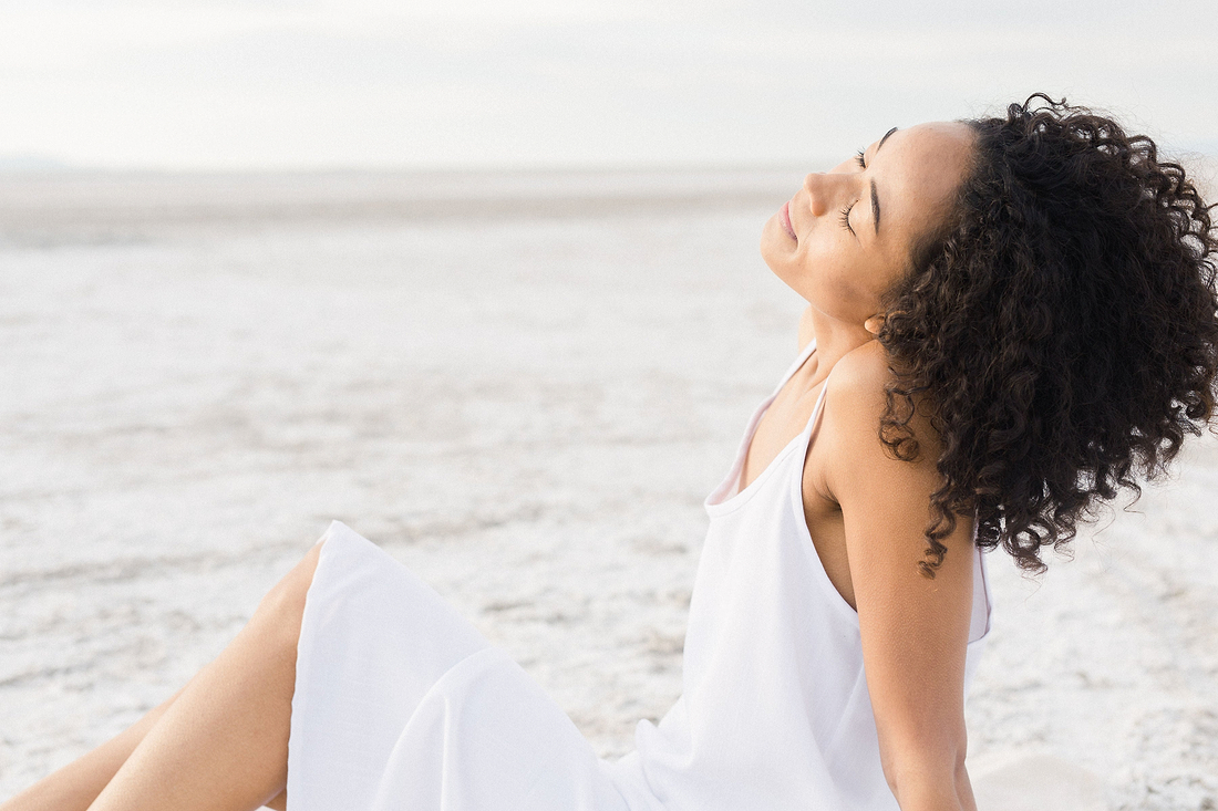 Woman in a white dress sitting outdoors on a bright salt flat with eyes closed and curly hair blowing back