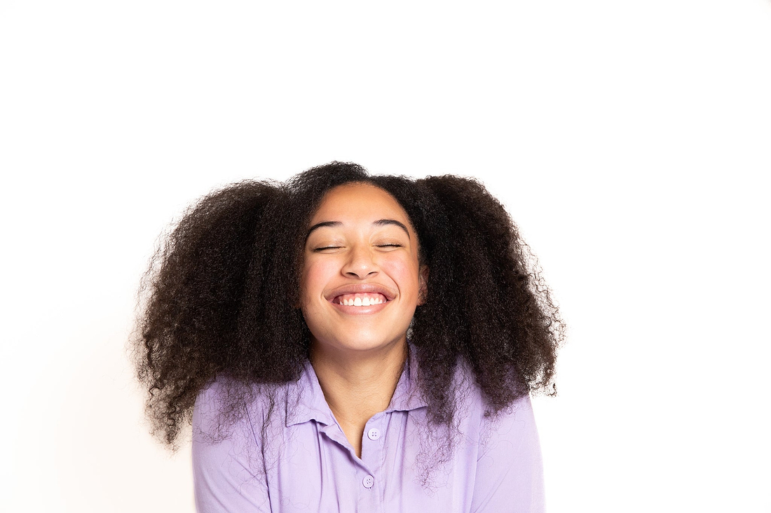 Smiling person with curly hair wearing a lavender shirt against a light background.