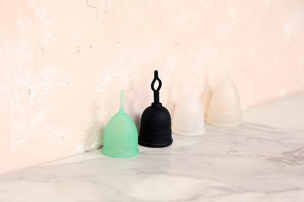 Reusable menstrual cups in various colors and styles displayed on a marble surface against a soft pink textured wall.