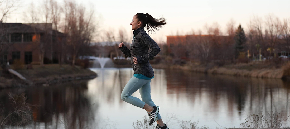 Woman jogging beside a calm lake at sunset wearing athletic clothes.