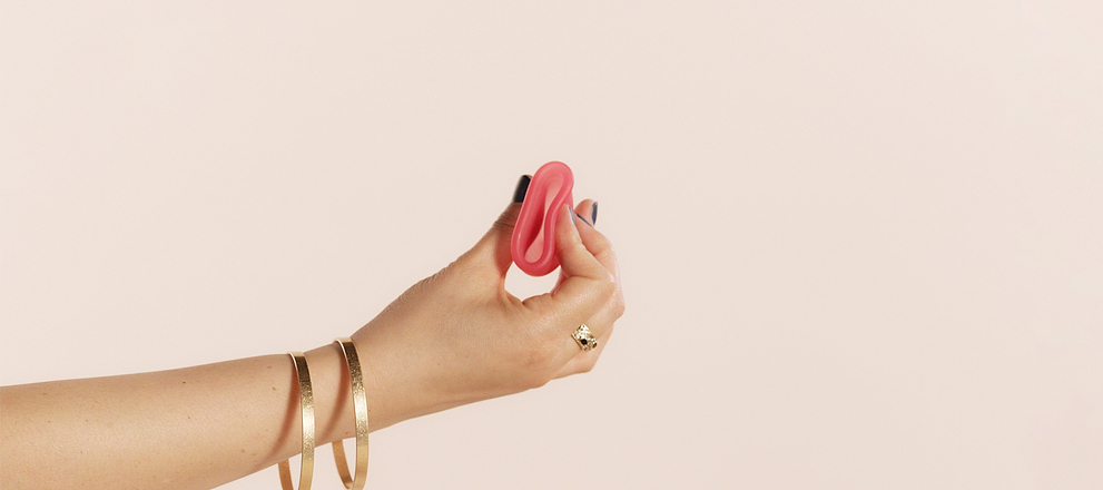 Hand holding a pink menstrual cup, wearing bracelets, against a neutral background.