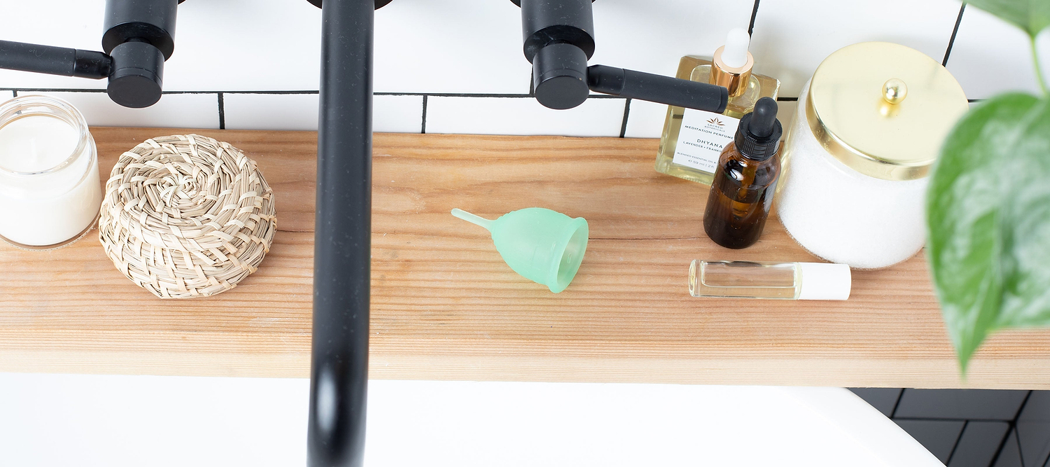 Green menstrual cup on a wooden bathroom shelf beside candles, oils, and toiletries.