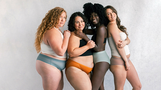 Four diverse women in colorful underwear and bras smiling together in a studio portrait against a neutral background.