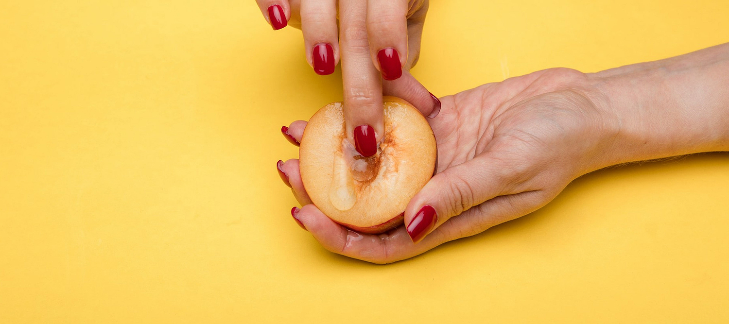 Hands with red nails holding a halved peach against a yellow background.