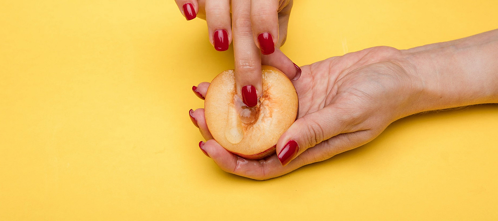 Hands with red nails holding a halved peach against a yellow background.