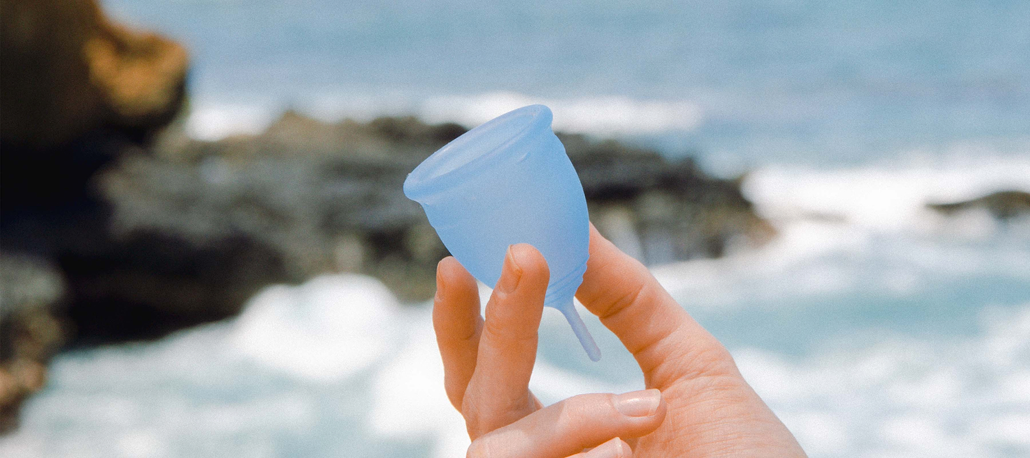 Hand holding a blue menstrual cup near ocean waves and rocky shoreline.