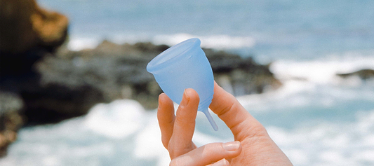 Hand holding a blue menstrual cup near ocean waves and rocky shoreline.