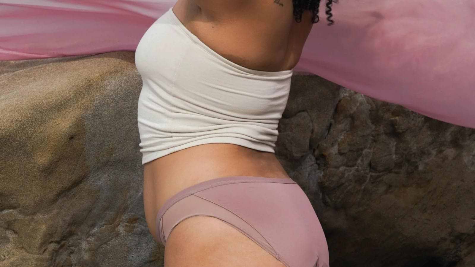 Person wearing light-colored underwear and tube top beside rocky background with pink fabric overhead.