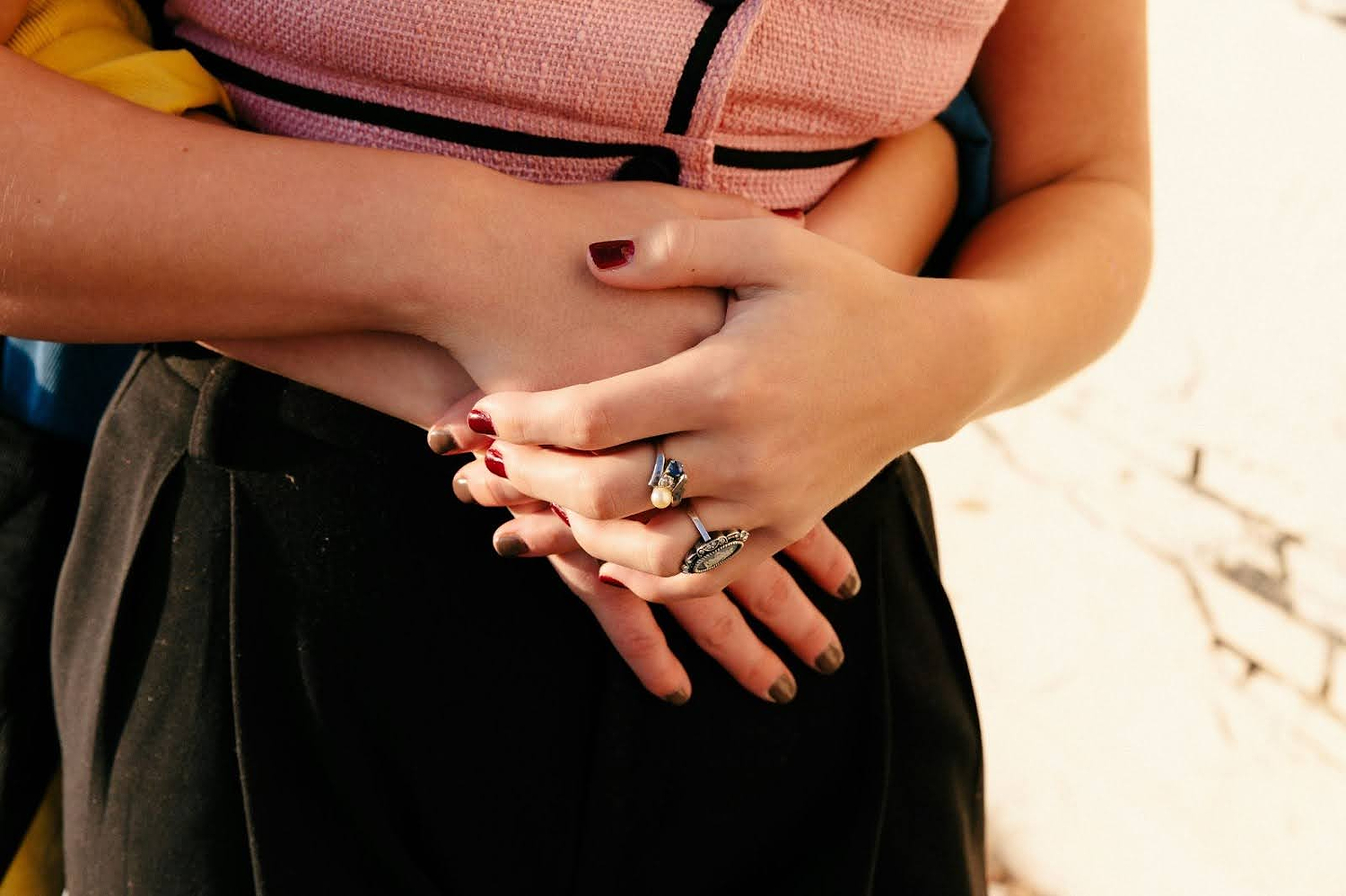 Person embracing another from behind, with clasped hands and rings visible, wearing pink and black clothing.