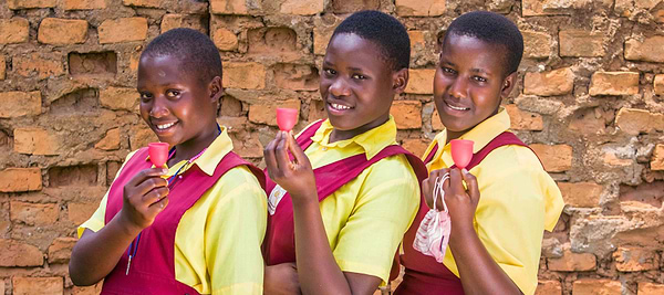 Three students holding menstrual cups and smiling in front of a brick wall.