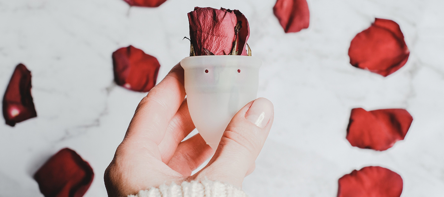 Hand holding a silicone menstrual cup with a dried rose inside, surrounded by scattered rose petals.